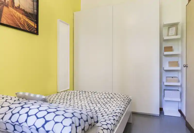 Chambre lumineuse et compacte avec couette à motifs, grand placard blanc et mur jaune chaleureux — parfaite pour un séjour cosy en solo ou en couple.