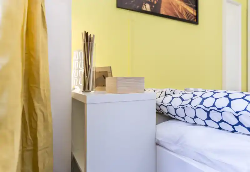 Bright, modern bedroom corner with a bedside table, decorative lamp and a patterned duvet partially visible — cozy, well-lit and styled for rental photos.