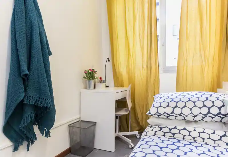 Coin chambre lumineux et fonctionnel avec literie à motifs, petit bureau près de la fenêtre et rideaux jaunes apportant une atmosphère chaleureuse.