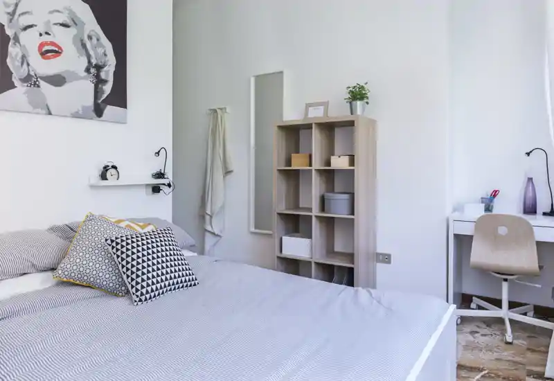Camera da letto moderna e luminosa con un grande letto con biancheria a righe, cuscini decorativi e una mensola minimal; sulla destra un piccolo scrittoio per lavorare.