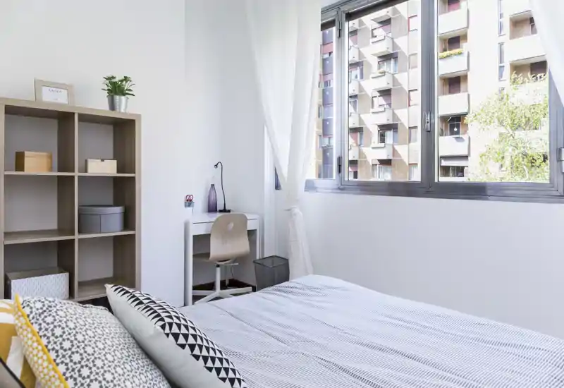 Camera da letto luminosa e moderna con letto ben rifatto, cuscini decorativi e una piccola scrivania vicino a una grande finestra che offre molta luce naturale.