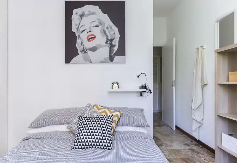 Camera da letto luminosa e moderna con letto matrimoniale ben rifatto, cuscini decorativi e una stampa d'arte sopra la testata. Linee pulite e luce naturale rendono l'ambiente accogliente e adatto per affitti.