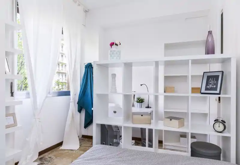 Angolo di camera luminoso e moderno con un letto basso in primo piano, una libreria bianca a giorno come divisorio e morbida luce naturale che filtra dalle tende leggere.