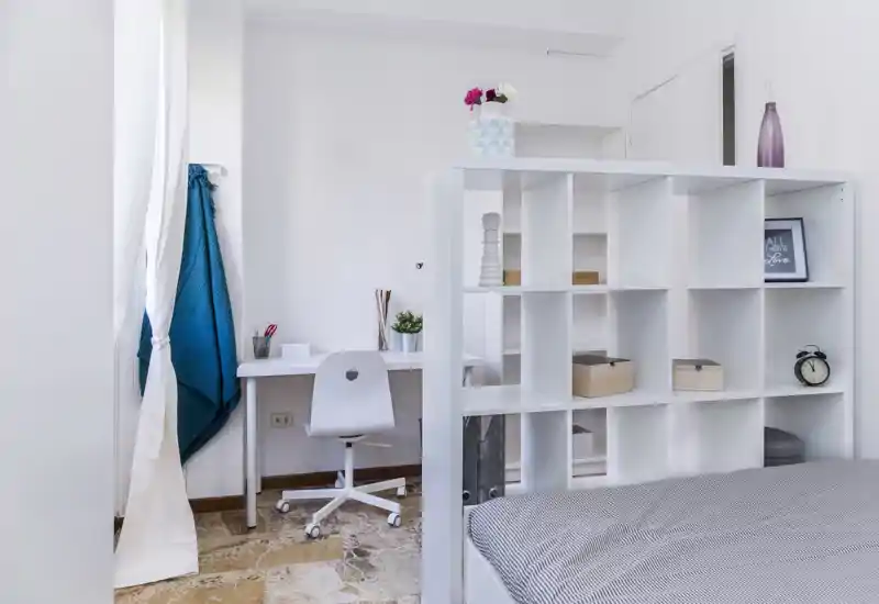 Camera da letto moderna e luminosa con letto basso parzialmente separato da un mobile a cubi bianco; una scrivania compatta e una sedia rendono lo spazio funzionale per lavoro e riposo.