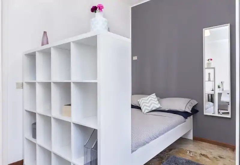 Camera da letto singola moderna in angolo, con letto ordinato dietro un divisorio a scaffali bianchi, cuscino dal motivo morbido e specchio a figura intera che riflette la stanza. Luminoso, minimal e ben curato — ideale per annunci che valorizzano un'area notte compatta e organizzata.
