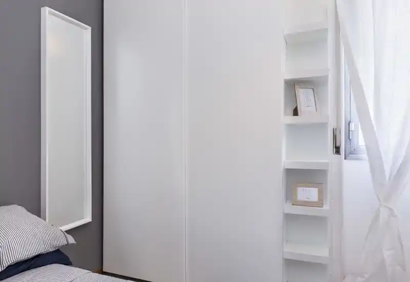 Angolo di camera minimal e luminoso con cuscino rigato e parte del materasso visibili, uno specchio alto a parete, scaffalature integrate e un armadio bianco accanto a una finestra con tenda leggera.