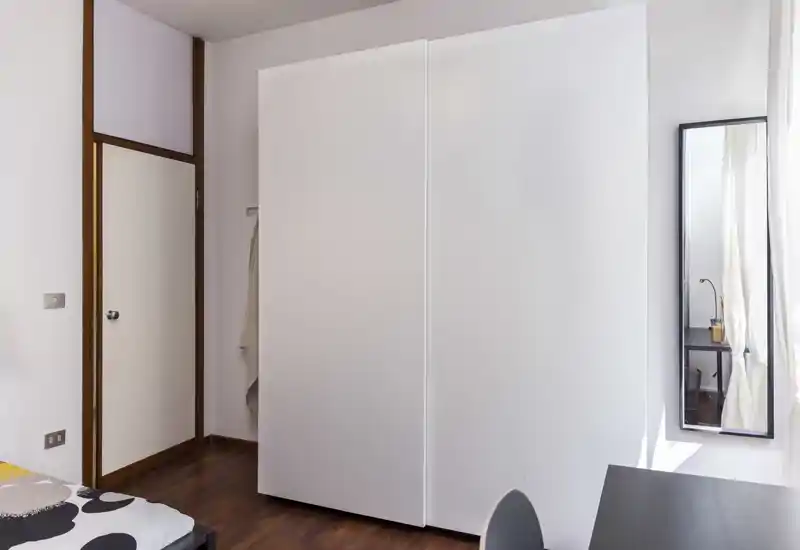 Rincón de dormitorio moderno y luminoso con un gran armario blanco, borde de la cama con ropa de cama estampada, espejo de pared y un pequeño escritorio — ordenado y bien iluminado.