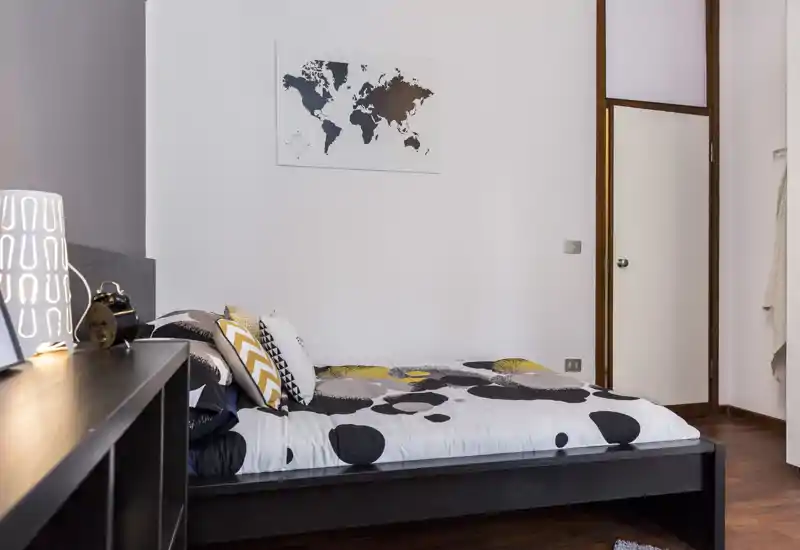 Dormitorio moderno y luminoso con edredón estampado, cojines decorativos y cama baja de madera; un cuadro sencillo y una lámpara de mesilla crean un ambiente acogedor y bien presentado.