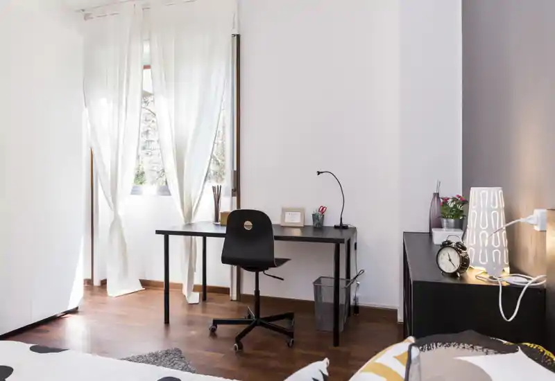 Rincón de dormitorio luminoso y moderno con cama en primer plano, escritorio junto a la ventana y decoración cuidada (lámpara y planta) — ideal para descansar y trabajar.