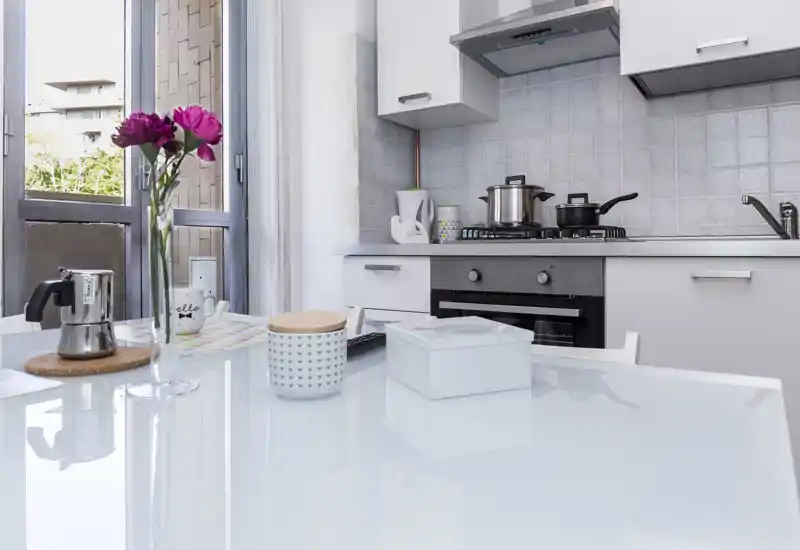 Bright modern kitchen with a glass-topped dining surface, stovetop and oven, decorative jar and fresh flowers that give a welcoming feel.