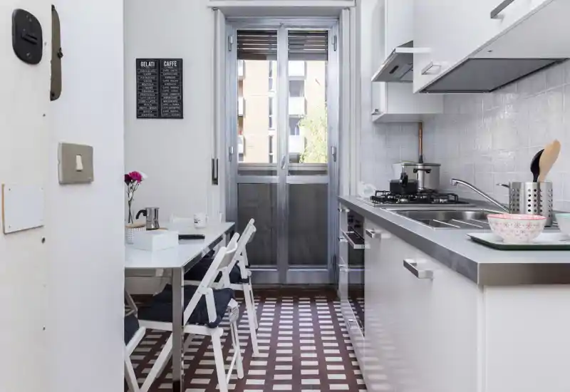 Compact, modern kitchen with a stainless sink, gas hob and a small dining table by a glass door leading to a balcony; clean white cabinets and patterned floor tiles create a bright, stylish space.