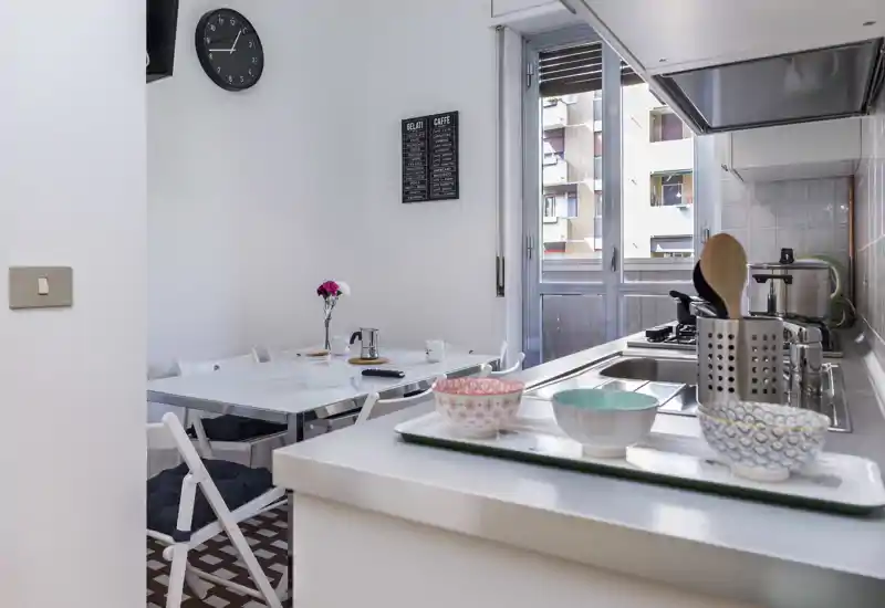 Bright, modern kitchen with a breakfast bar and a small dining area. Natural light from the window highlights tidy countertops, colorful bowls and a cozy table setting.