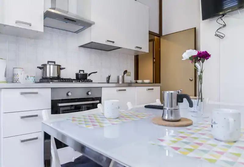 Bright, modern kitchenette with a gas stove, oven, white cabinets and a small dining table set with cups and a moka pot — tidy and inviting for short stays.