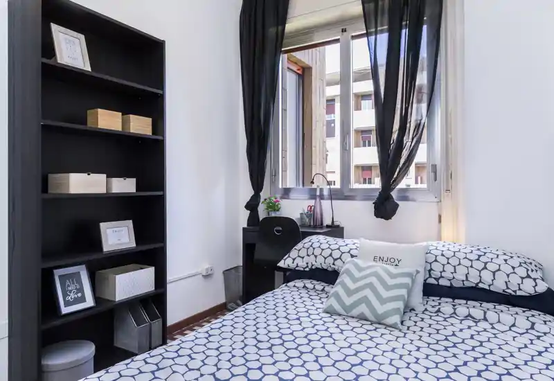 Chambre lumineuse et bien mise en scène avec linge de lit à motifs, coussins décoratifs et un petit bureau près de la fenêtre — idéale pour une personne ou un couple.