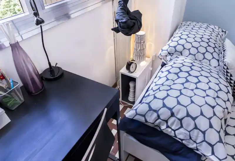 Coin chambre compact avec un lit simple habillé de linge à motif géométrique, une petite étagère de chevet avec lampe et réveil, et un bureau près de la fenêtre—lumineux, propre et chaleureux.