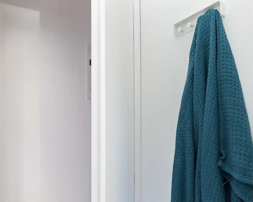 A minimal entrance/coat area with a teal textured towel or light jacket hanging on a wall hook against a white doorframe.
