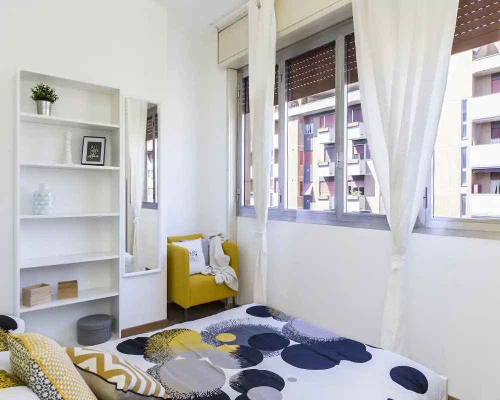 Bright, modern bedroom with a patterned bedspread, large windows drenching the space in natural light, and a cozy yellow armchair by the window.