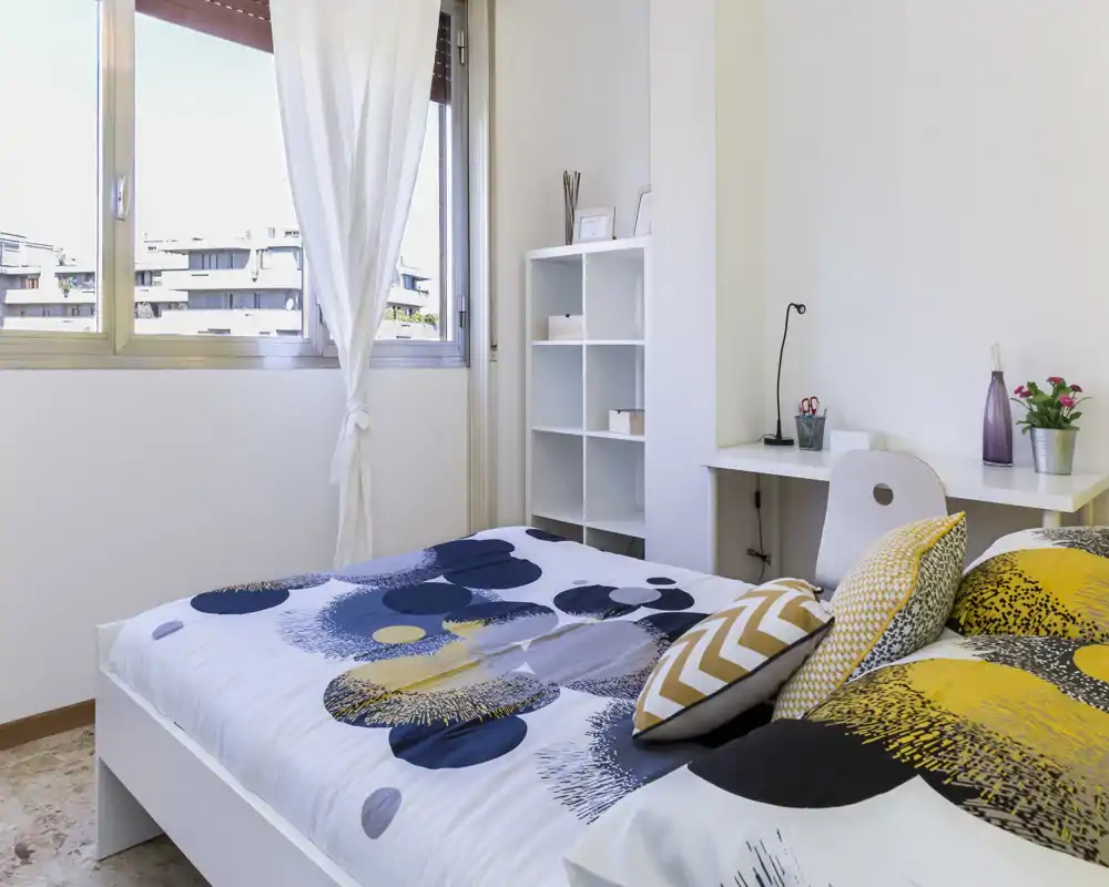 Bright, modern bedroom with patterned bedding, decorative cushions and a workspace by the wall. Large window provides plenty of natural light and a clean, minimal shelving unit adds functional storage.