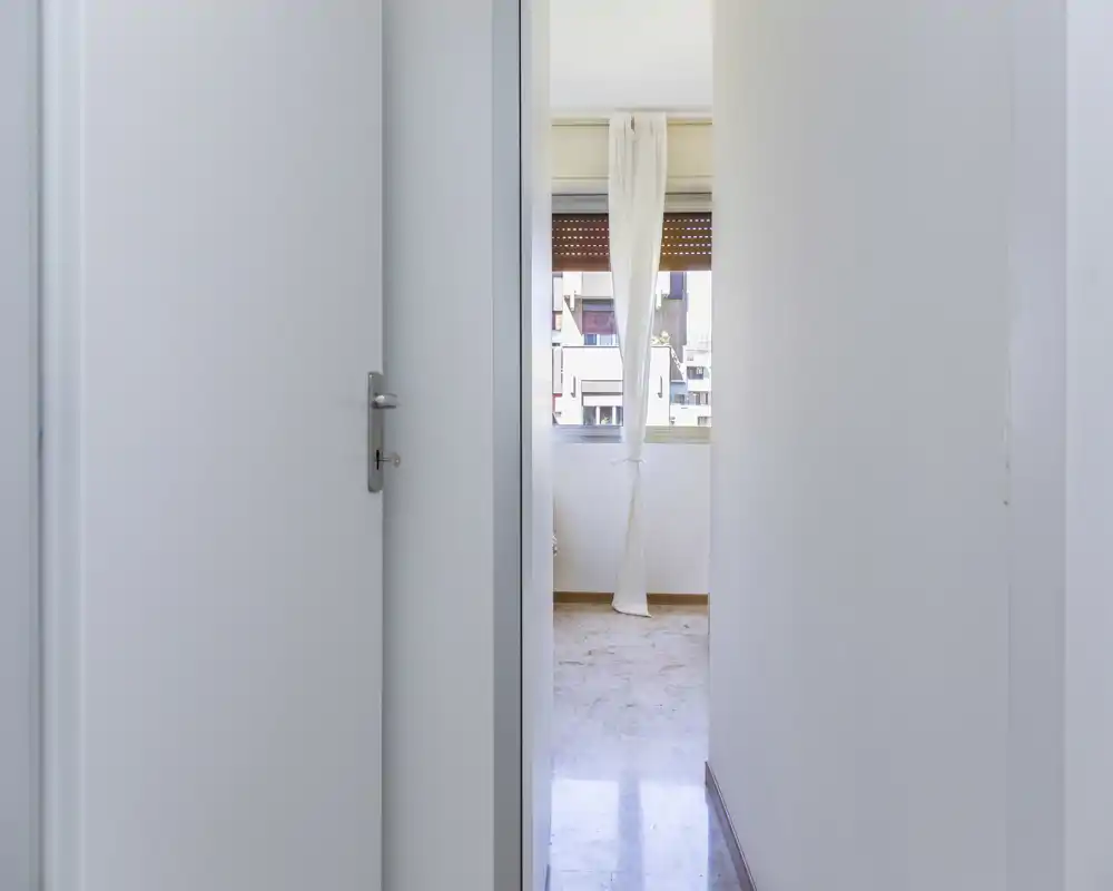 Narrow, bright hallway/entrance leading to a windowed room; clean white walls and a simple curtain allow natural light to flow through.