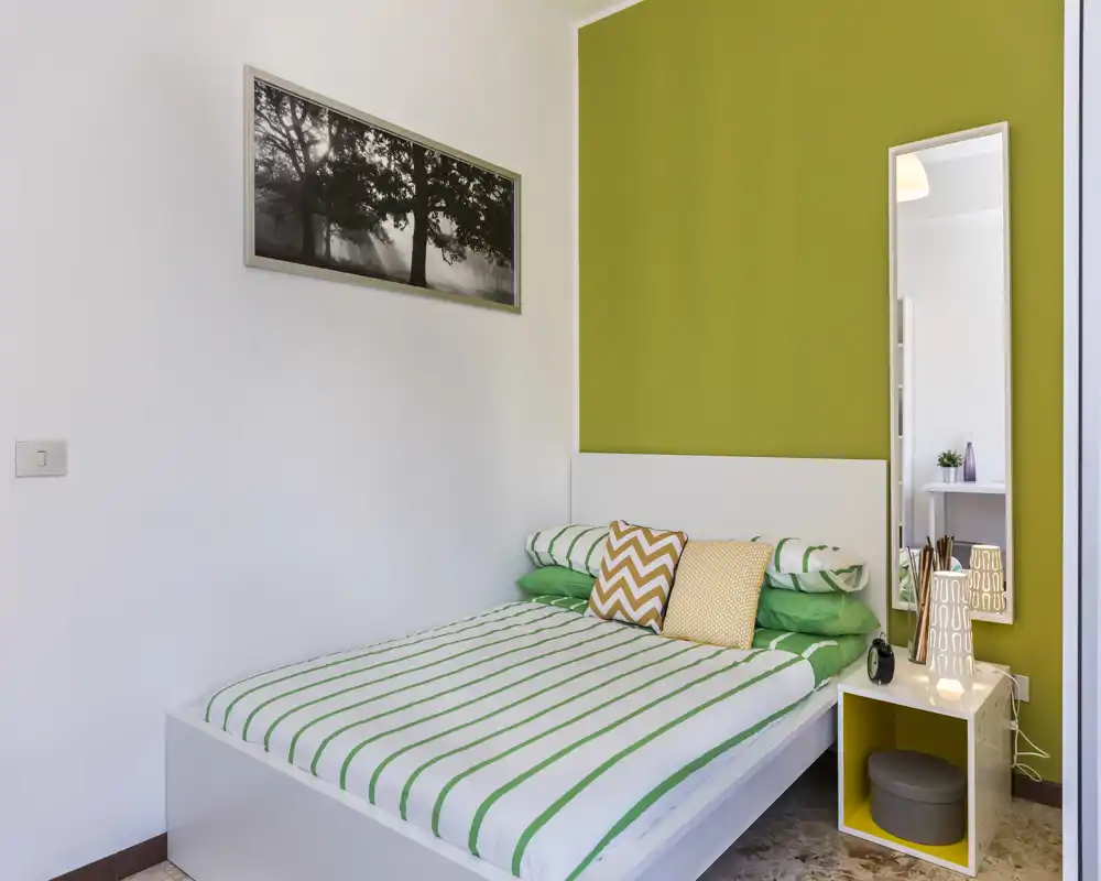 Bright, modern bedroom with a green-accent wall and a neatly made double bed featuring striped linens and decorative cushions. A tall mirror and minimalist bedside table complete the fresh, welcoming layout ideal for rental photos.