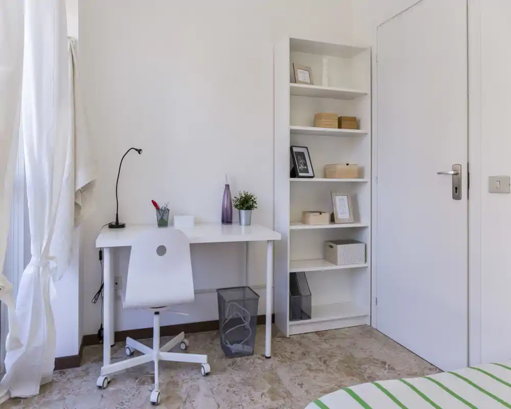 Bright, minimalist bedroom corner with a white desk, shelving and a small visible portion of the bed; ideal as a study nook within the sleeping area.
