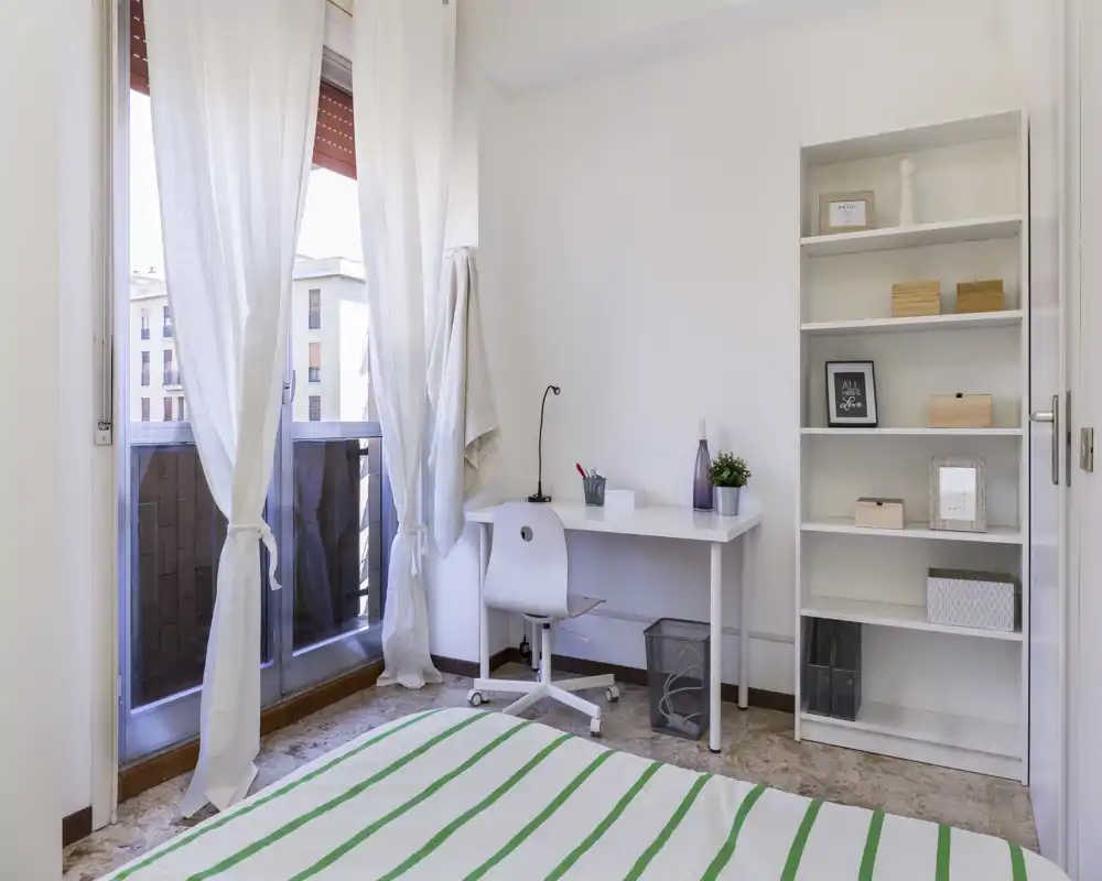Bright, minimalist bedroom with a white desk and shelving next to a balcony; striped bedspread and airy curtains create a fresh, uncluttered atmosphere ideal for short-term rentals.