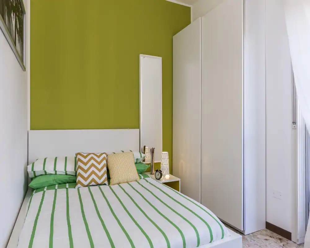 Bright, modern bedroom with a striped green-and-white bed, decorative cushions and a tall wardrobe — clean, well-lit space ideal for relaxing.