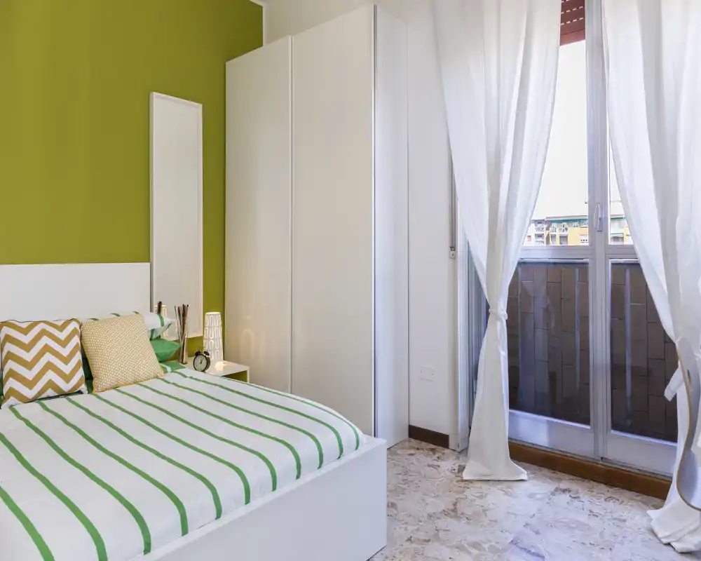 Bright, modern bedroom with a white bed dressed in green-striped linens, decorative cushions and a bedside table lamp; large window with sheer curtains provides abundant natural light.