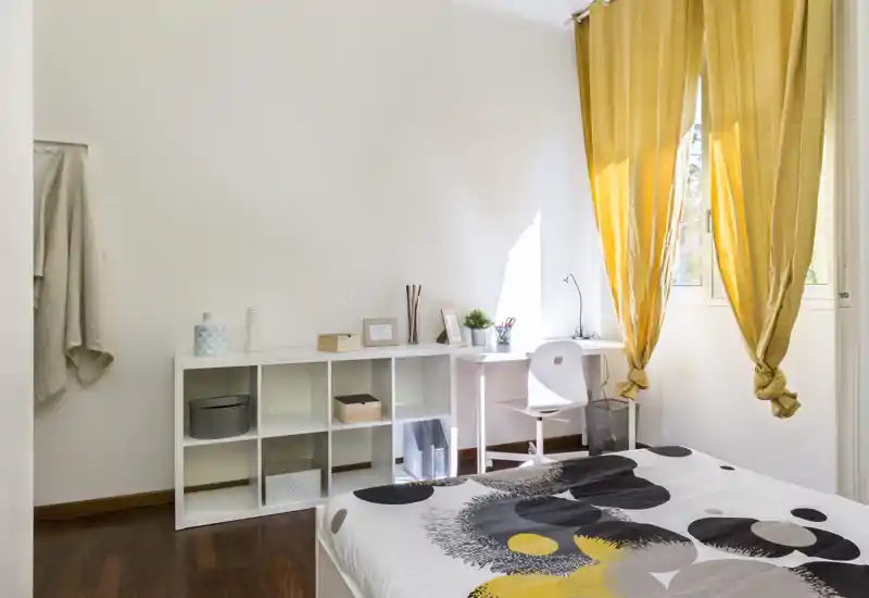 Dormitorio luminoso y bien decorado con edredón estampado, estantería blanca y un pequeño escritorio junto a la ventana; las cortinas amarillas aportan calidez y la luz natural llena la habitación.