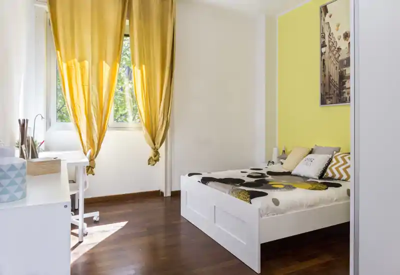 Dormitorio luminoso y soleado con cama individual blanca, ropa de cama estampada y toques amarillos cálidos; escritorio junto a la ventana para un rincón de estudio claro.