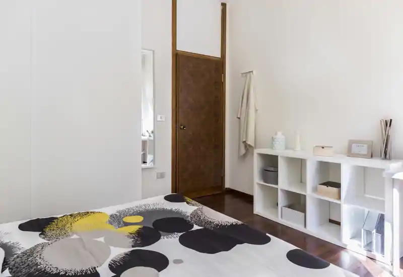 Dormitorio moderno y luminoso con edredón estampado, cama baja en primer plano y estantería blanca de cubos en la pared; suelo de madera y luz natural que aportan una sensación cómoda y aireada.