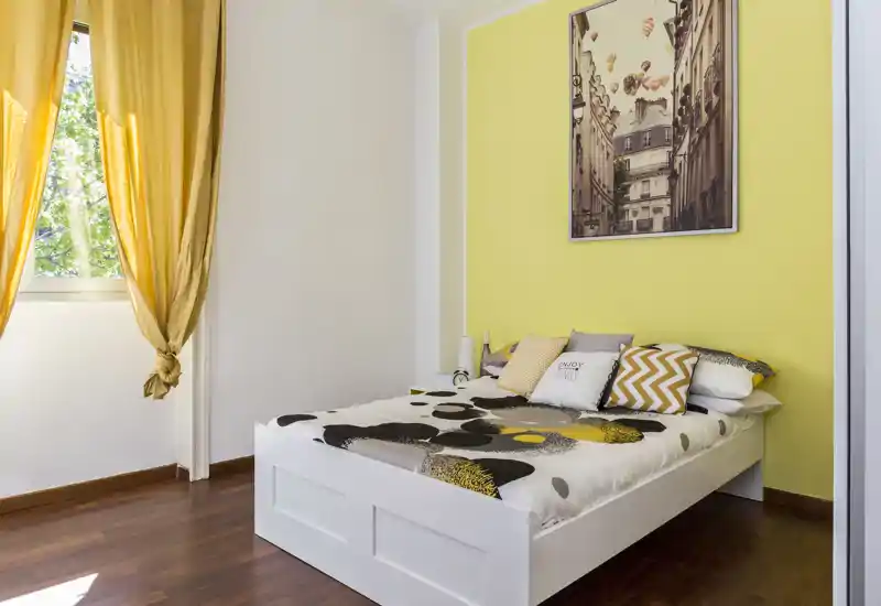 Dormitorio moderno y luminoso con cama blanca de madera, ropa de cama con estampado y detalles amarillos coordinados; ventana grande con cortinas que aporta luz natural.