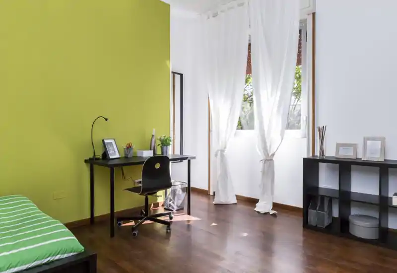 Camera da letto luminosa e moderna con parete verde a contrasto, letto singolo con copriletto a righe, scrivania compatta e scaffale — ideale per uno studente o una persona sola.