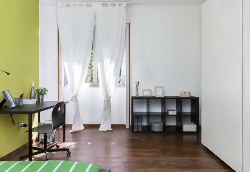 Angolo di camera da letto luminoso e minimal con una piccola porzione del letto visibile, una scrivania vicino a una parete verde e scaffalatura per riporre oggetti. La luce naturale attraversa le tende bianche donando un'atmosfera fresca e ariosa.