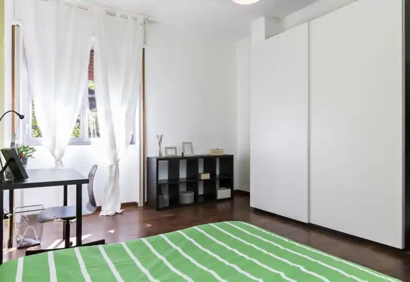 Camera da letto luminosa e minimalista con letto rifatto in primo piano, grande armadio bianco e una piccola scrivania vicino alla finestra che lascia entrare luce naturale.