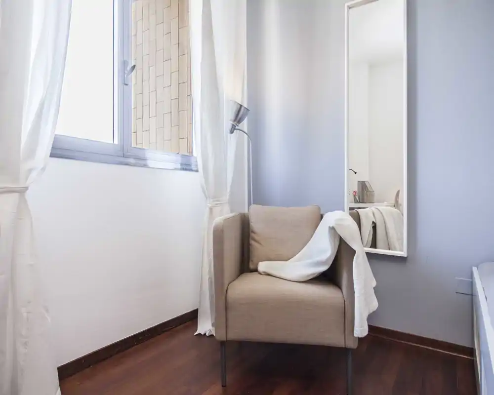 Pequeña zona de estar con un puf/sillón claro junto a una ventana luminosa; textiles suaves aportan calidez pese a las anomalías de la imagen.
