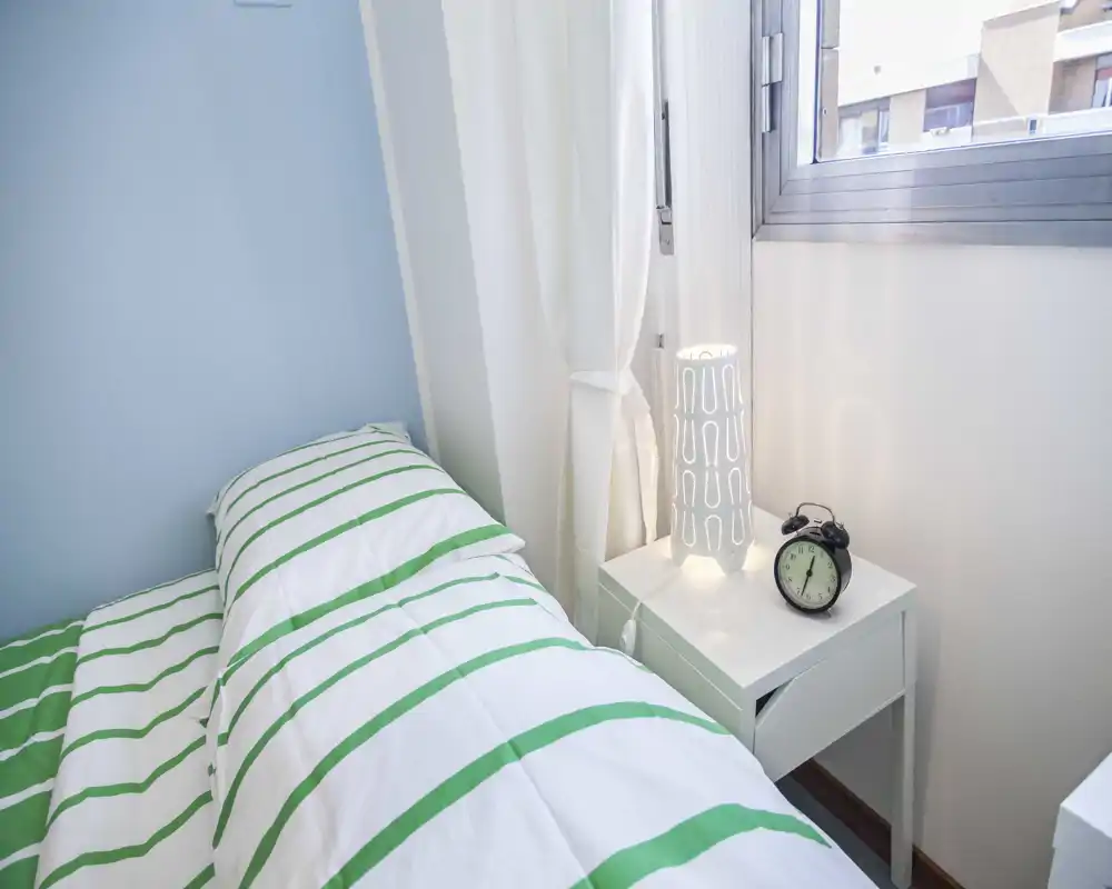Camera da letto compatta e luminosa con cuscini e comodino; l'immagine presenta artefatti visivi ma mostra un'area notte accogliente dai toni neutri.
