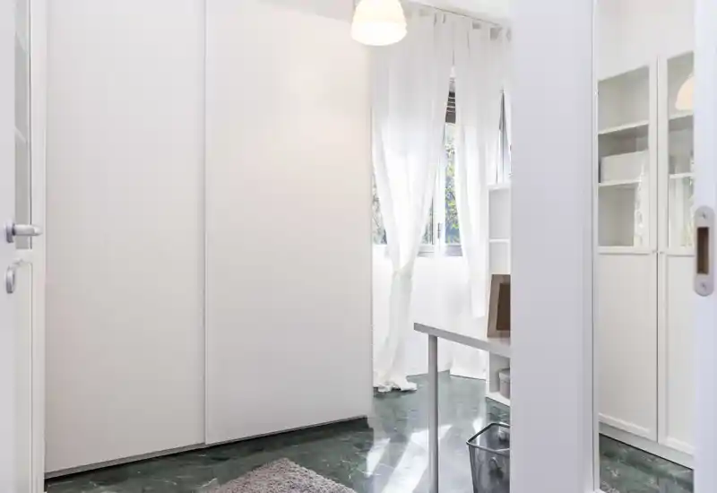 Angolo di camera minimalista con armadi bianchi, tende leggere e luce naturale; pavimento in marmo e tappeto creano un'area notte calma e neutra.
