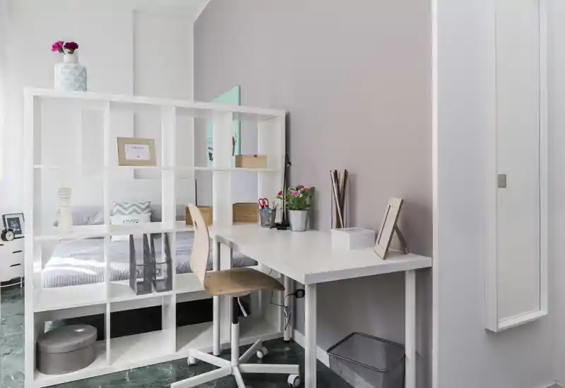 Camera da letto luminosa e ben arredata con area studio: una libreria bianca divide il letto ordinato dalla scrivania compatta e dalla sedia, cuscini decorativi e piante aggiungono un tocco accogliente.