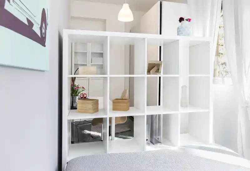 Camera da letto moderna e luminosa con un letto a righe in primo piano e una libreria a cubi bianca che divide lo spazio; la luce naturale attraversa la finestra con tende creando un’atmosfera ariosa e pulita.