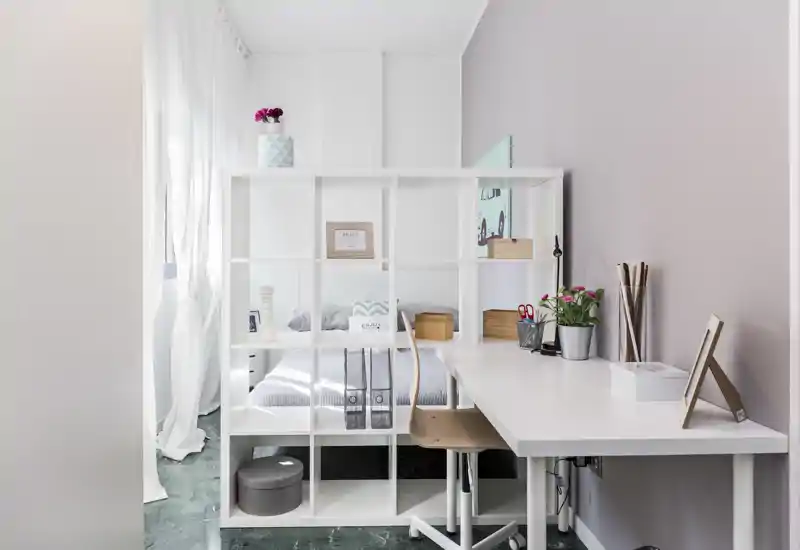 Camera da letto moderna e luminosa con una libreria a vista che separa il letto da una scrivania lunga; tessili neutri e piccoli accessori decorativi rendono l'ambiente accogliente.