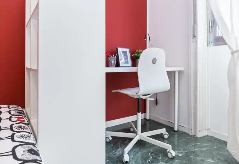 Small bedroom corner with a compact desk and swivel chair beside a partially visible bed; bright window light and red accent wall give the space a fresh, modern feel.