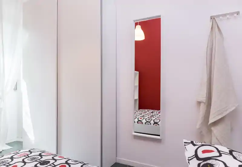 Small, bright bedroom corner with patterned bedding, a tall mirror reflecting a red accent wall, and a built-in wardrobe. The photo emphasizes brightness and a tidy, modern style.