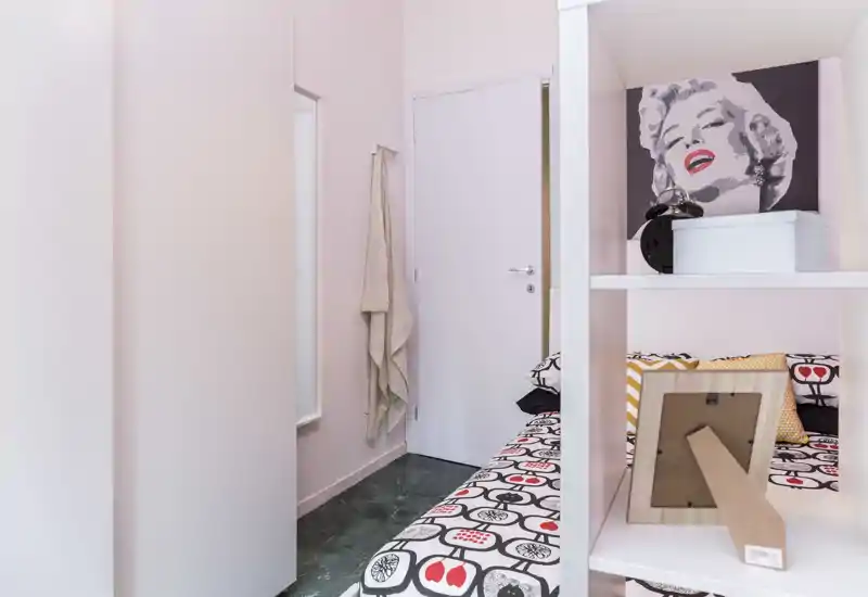 Camera da letto compatta e ben arredata con biancheria a motivo, cuscini decorativi e quadro pop-art; luminosa, ordinata e accogliente, letto singolo/doppio vicino a scaffalatura.