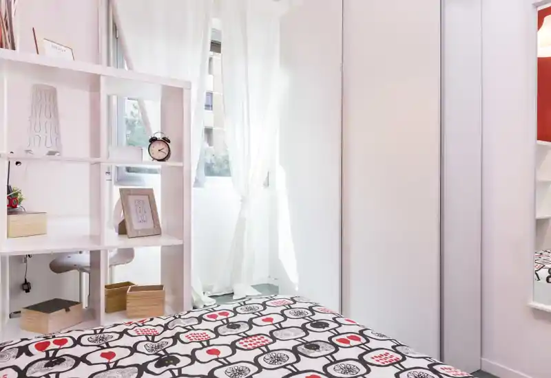 Camera da letto moderna e luminosa con copriletto a motivi, scaffalatura bianca e una finestra che lascia entrare luce naturale. Linee pulite e arredo semplice rendono l'ambiente accogliente.