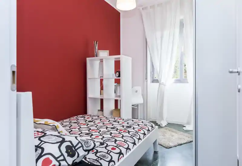 Camera da letto luminosa e ordinata con parete rossa a contrasto, biancheria con motivo, libreria a cubi e finestra che illumina la stanza — accogliente e moderna.