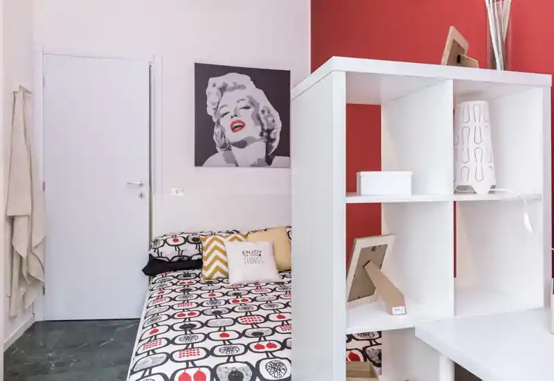 Camera da letto luminosa e di stile con copriletto a fantasia, cuscini decorativi e una libreria bianca che divide parzialmente lo spazio. La parete rossa e l'opera di Marilyn donano personalità e un tocco moderno.