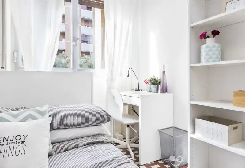 Dormitorio luminoso y estiloso con cama bien hecha, pequeño escritorio blanco junto a la ventana y estanterías de buen gusto — ideal para un alquiler de estudiante o individual.