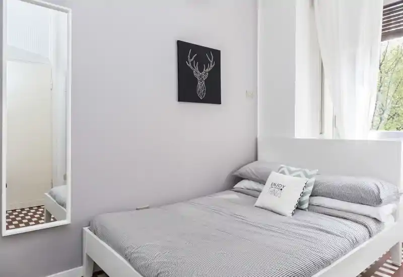 Dormitorio luminoso y minimalista con cama doble bien hecha, ropa de cama gris y cojines decorativos junto a una ventana que aporta luz natural.