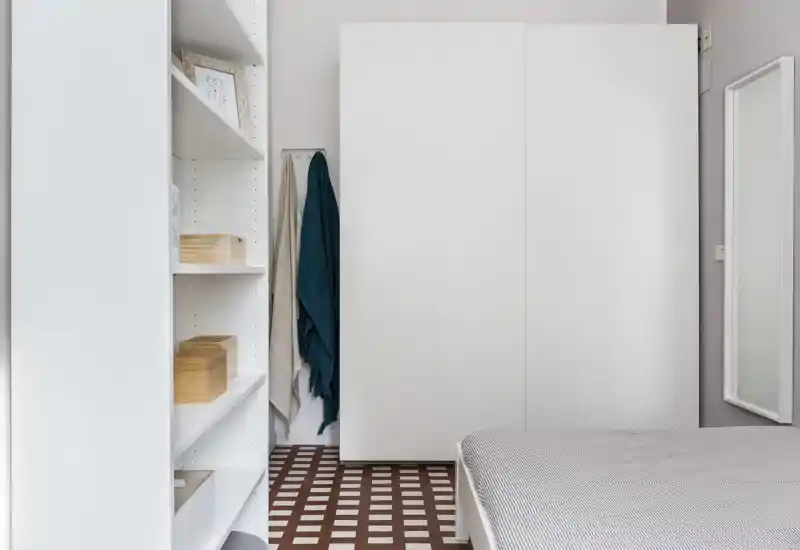 Rincón de dormitorio minimalista y bien iluminado con cama parcialmente visible, gran armario blanco, estanterías y suelo de baldosas con patrón; estilo contemporáneo y sereno.